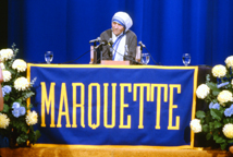 Mother Teresa seated at table with Marquette banner